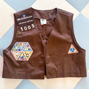 May include: Brown Girl Scouts USA vest with patches. The vest has patches that read "Girl Scouts USA", "Michigan Metro", and "1005". There are also several colorful patches with various designs. The vest is made of a smooth fabric.