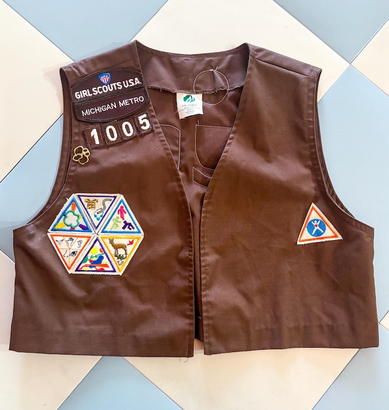 May include: Brown Girl Scouts USA vest with patches. The vest has patches that read "Girl Scouts USA", "Michigan Metro", and "1005". There are also several colorful patches with various designs. The vest is made of a smooth fabric.