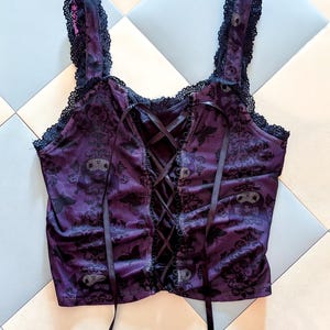 May include: A purple corset top with black lace trim and straps. The top features a black floral pattern and a lace-up back with black ribbon. The straps are trimmed with black lace. The top is made of a sheer fabric.