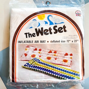 Puede incluir: Un colchón de aire inflable vintage de "The Wet Set" en su embalaje original. El embalaje presenta el nombre del producto, un gráfico de sol y olas, y las palabras "INFLATABLE AIR MAT desinflado tamaño 183 cm x 69 cm". El colchón se muestra con un diseño de corazón rojo y sandía.