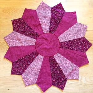 Peut inclure: Un dessous de plat matelassé fait à la main en forme d'étoile, dans des tons de rose et de violet. Le centre est un cercle rose foncé uni, entouré de sections alternées de rose uni, d'imprimé floral et de tissu rose plus clair. Le design est symétrique et détaillé.