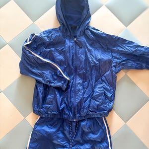 May include: A blue athletic tracksuit set, featuring a hooded jacket with white stripes on the sleeves and matching pants. The jacket has a zipper closure. The drawstring pants have white stripes down the sides.