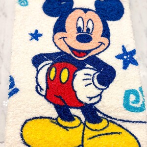 May include: A rectangular, off-white rug featuring a smiling Mickey Mouse character. Mickey is depicted in his classic colors: blue ears and shorts, red shorts, yellow shoes, and white gloves. Blue stars and swirls decorate the background.