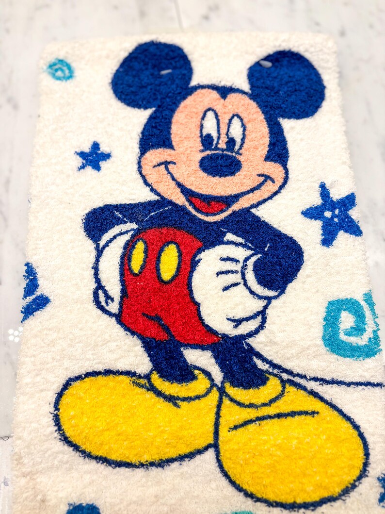 May include: A rectangular, off-white rug featuring a smiling Mickey Mouse character. Mickey is depicted in his classic colors: blue ears and shorts, red shorts, yellow shoes, and white gloves. Blue stars and swirls decorate the background.