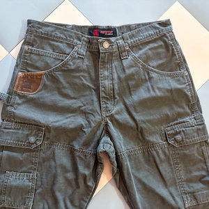 May include: Olive green cargo pants with multiple pockets and a brown leather patch detail. The pants have a button and zipper closure, belt loops, and a classic five-pocket design. The pants are made of a durable fabric.