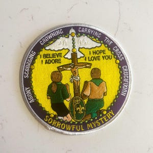 May include: A circular patch with a yellow center and a purple border. The patch features two kneeling figures, a cross, and the text "SORROWFUL MYSTERY." Additional text includes "CARRYING THE CROSS" and "CRUCIFIXION."