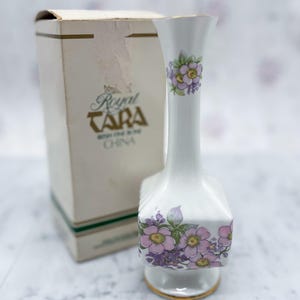 May include: A white porcelain vase with a floral design and gold trim, next to a box with the words "Royal Tara" printed on it. The vase features pink and purple flowers. The box is beige with green and gold accents.
