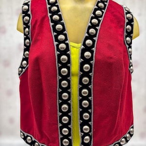 May include: A red vest with black trim and silver-colored studs. The vest has a V-neck and is sleeveless. The trim is along the edges and around the neckline. A yellow top is visible underneath the vest.