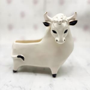 Puede incluir: Un macetero de cerámica con forma de toro en blanco roto con detalles negros. El toro tiene una cara detallada, pequeños cuernos y una abertura rectangular para plantas. Un elemento decorativo para el hogar.
