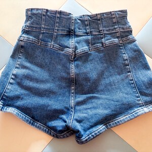 May include: High-waisted denim shorts in a medium wash blue. The shorts feature a structured waistband with multiple vertical seams and a classic five-pocket design. The back view shows the shorts' tailored fit and the detailed stitching.