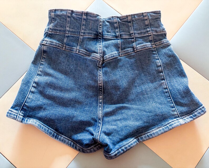 May include: High-waisted denim shorts in a medium wash blue. The shorts feature a structured waistband with multiple vertical seams and a classic five-pocket design. The back view shows the shorts' tailored fit and the detailed stitching.