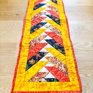May include: A colorful, rectangular quilted table runner. The runner features a patchwork design with yellow, red, white, and navy blue triangles. The edges are trimmed with a yellow border and a red binding.