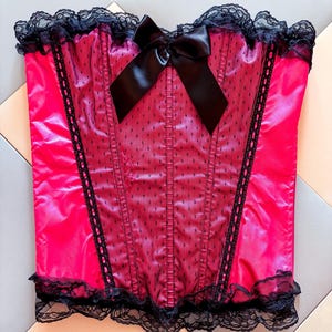 May include: A vibrant pink corset with black lace trim and a satin bow. The corset features a dotted mesh overlay on the front panel and vertical boning details. The top and bottom edges are finished with delicate black lace.
