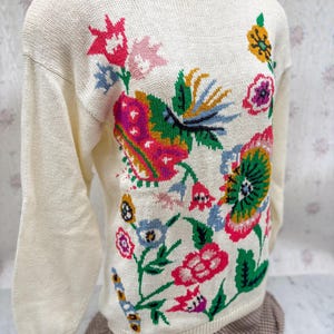 May include: Cream-colored sweater with a crew neck and long sleeves. The sweater features a vibrant floral design in pink, green, yellow, and blue. The flowers are intricately detailed, creating a colorful and eye-catching pattern.