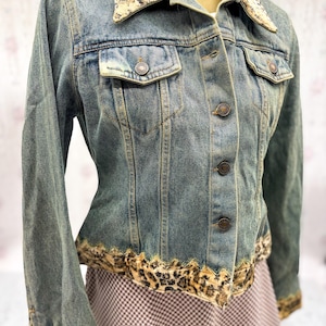 May include: A blue denim jacket with a leopard print collar and trim. The jacket has button fastenings, two chest pockets, and long sleeves. The leopard print provides a striking contrast to the denim material.