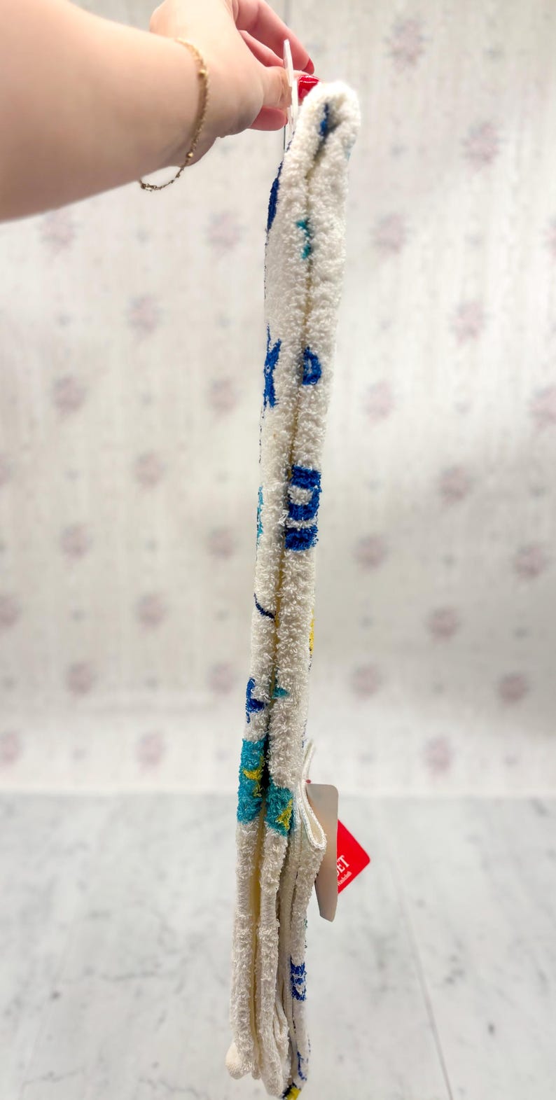 May include: White, fluffy socks with blue and yellow accents. The socks have a textured appearance and feature a pattern of blue letters and shapes. A red tag is attached to the socks.