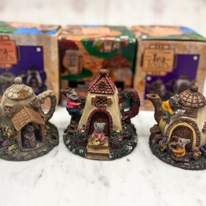 Könnte beinhalten: Drei dekorative Keramik-Teekannen mit hausähnlichen Designs. Jede Teekanne hat ein einzigartiges Farbschema und architektonische Details, darunter Türen, Fenster und Miniaturfiguren. Die Teekannen werden vor ihren Originalverpackungen ausgestellt.