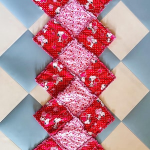 May include: A handmade, quilted table runner featuring a repeating pattern of red squares with Snoopy and heart designs, alternating with pink squares covered in tiny hearts. The edges have a frayed, textured finish.