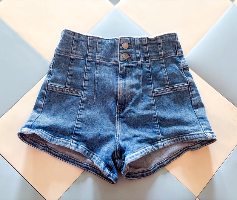 May include: High-waisted blue denim shorts with a button closure. The shorts feature a classic five-pocket design and a tailored fit. The denim has a medium wash and visible stitching details.