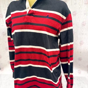 May include: A long-sleeved rugby shirt with horizontal stripes in red, black, and white. The shirt has a white collar and a three-button placket. A small blue logo is visible on the chest.