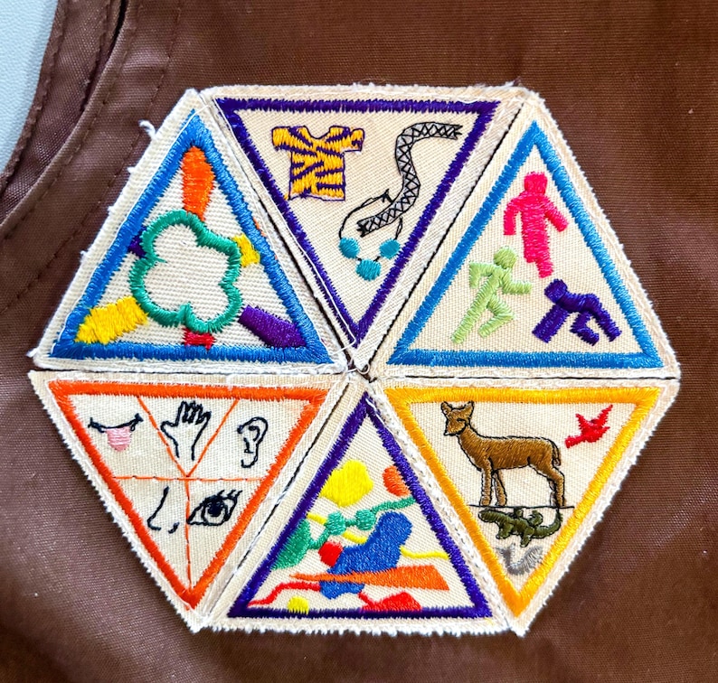 May include: A collection of colorful triangular patches with various embroidered designs. The patches feature images of a tree, a snake, running figures, a deer, and symbols representing senses. The patches are sewn onto a brown fabric.