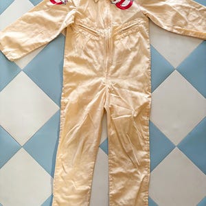 Könnte beinhalten: Beigefarbener Overall mit Kragen und Reißverschluss vorne. Zwei rot-weiße Ghostbusters-Logo-Aufnäher sind auf den Schultern angebracht. Zwei Reißverschlusstaschen sind auf der Brust sichtbar. Das Kleidungsstück liegt flach auf einer blau-weißen Fliesenoberfläche.