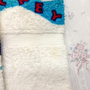May include: White terry cloth towel with a blue and turquoise design and red lettering. The towel has a white border and a soft, textured surface. The design includes the letters "CKEY".