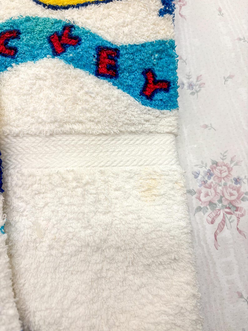 May include: White terry cloth towel with a blue and turquoise design and red lettering. The towel has a white border and a soft, textured surface. The design includes the letters "CKEY".