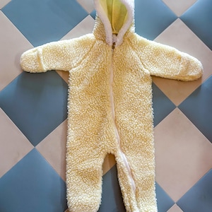 May include: A light yellow baby onesie with a hood and a full-length zipper. The hood is lined with white and light yellow fabric. The onesie has long sleeves and attached feet. The fabric has a textured, fuzzy appearance.