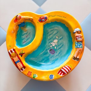 May include: A yellow ceramic bowl with a pool-themed design featuring two blue water sections, a person swimming, a person sunbathing on a red and white striped towel, a person reading a newspaper, a white dog with a ball, and the text Sum Sum Summertime.