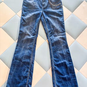 May include: A pair of blue denim jeans featuring a faded wash, a button and zipper fly closure, and a decorative metallic chain detail attached to the front right pocket area. The jeans have a straight-leg cut with visible stitching along the seams.