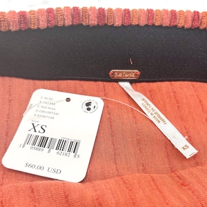 May include: Close-up of a coral-colored skirt with a ribbed texture. The waistband is black with a corduroy-like trim in shades of orange and red. A white tag indicates the size is XS.