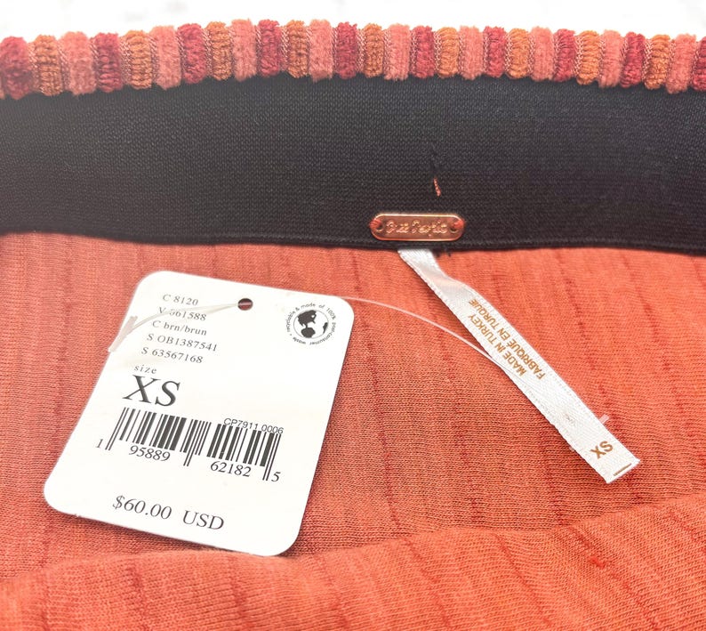 May include: Close-up of a coral-colored skirt with a ribbed texture. The waistband is black with a corduroy-like trim in shades of orange and red. A white tag indicates the size is XS.