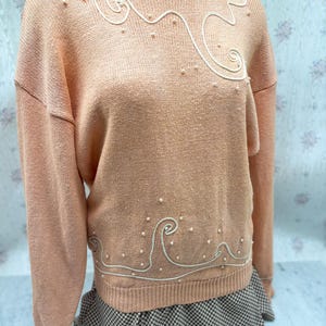 May include: A peach-colored sweater with a crew neck and long sleeves. The sweater features a white, wave-like design with small, pearl-like embellishments. The bottom hem and cuffs are ribbed. The sweater is displayed on a mannequin.