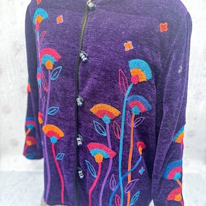 May include: A purple jacket with a floral design. The jacket features colorful embroidered flowers in shades of orange, blue, and pink. It has a round neckline, long sleeves, and a button-up front. The jacket's trim is black.