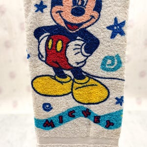 May include: White hand towel featuring a colorful embroidered image of Mickey Mouse. Mickey is depicted in his classic outfit with yellow shoes, red shorts, and white gloves. The word "MICKEY" is embroidered in blue letters.