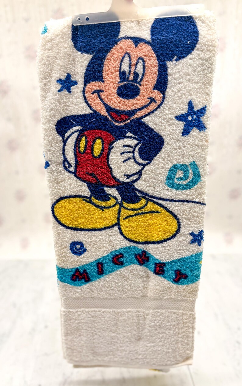 May include: White hand towel featuring a colorful embroidered image of Mickey Mouse. Mickey is depicted in his classic outfit with yellow shoes, red shorts, and white gloves. The word "MICKEY" is embroidered in blue letters.