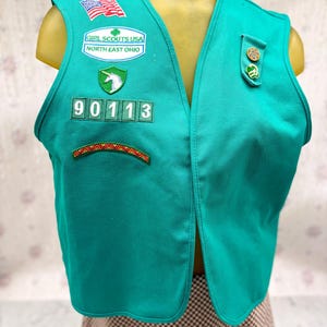 May include: Teal Girl Scouts USA vest with patches, including an American flag, a unicorn, and the text "Girl Scouts USA North East Ohio." The vest has the number "90113" and a decorative patch at the bottom. The vest is displayed on a mannequin.