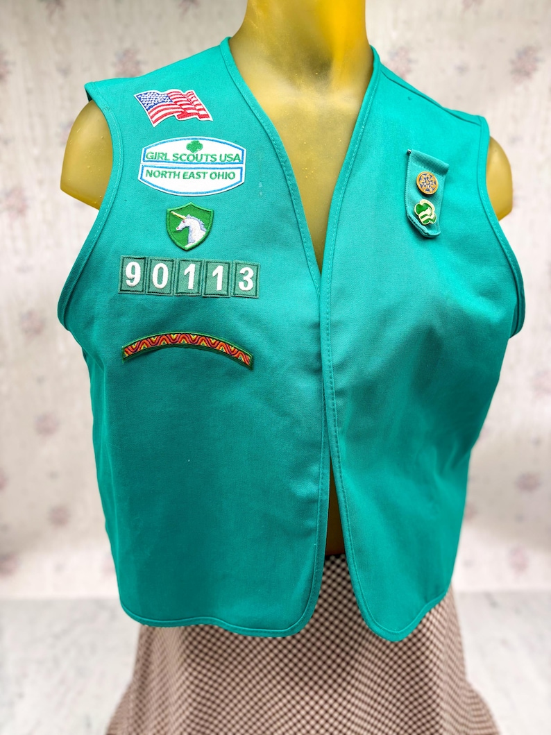 May include: Teal Girl Scouts USA vest with patches, including an American flag, a unicorn, and the text "Girl Scouts USA North East Ohio." The vest has the number "90113" and a decorative patch at the bottom. The vest is displayed on a mannequin.