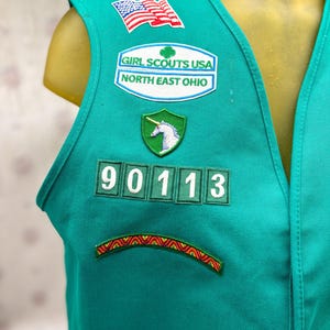 May include: Teal Girl Scouts vest with multiple patches. Patches include an American flag, "Girl Scouts USA North East Ohio", a unicorn, and the number "90113". A colorful embroidered design is at the bottom.