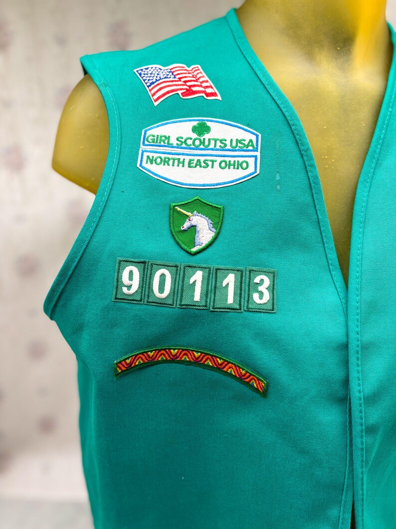 May include: Teal Girl Scouts vest with multiple patches. Patches include an American flag, "Girl Scouts USA North East Ohio", a unicorn, and the number "90113". A colorful embroidered design is at the bottom.