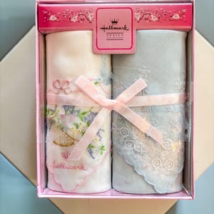 May include: A Hallmark Design Collection gift box containing two rolled handkerchiefs. One is white with floral embroidery and the other is light blue with lace trim. Both are tied with a pink ribbon. The box is pink with floral accents.