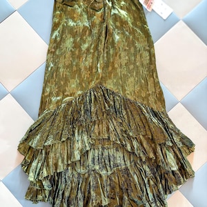 May include: Olive green maxi skirt with a floral pattern and tiered ruffles. The skirt has a drawstring waist and a flowing silhouette. The fabric has a slight sheen, giving it a luxurious look. The skirt is displayed on a checkered tile floor.