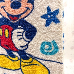 May include: White terry cloth towel featuring a colorful image of Mickey Mouse. Mickey is depicted with a blue outline, red shorts, yellow shoes, and white gloves. The towel also has blue star and swirl accents.