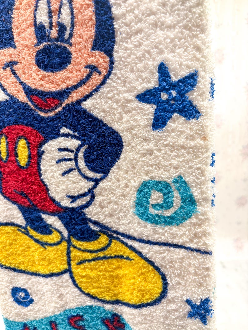May include: White terry cloth towel featuring a colorful image of Mickey Mouse. Mickey is depicted with a blue outline, red shorts, yellow shoes, and white gloves. The towel also has blue star and swirl accents.