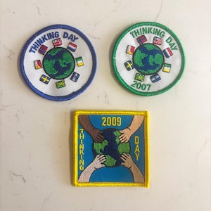 May include: Three embroidered patches. Two are circular with "Thinking Day" text and a globe surrounded by flags. One is blue and the other green, both with the year. The third is square, with hands around a globe and "Thinking Day" text.
