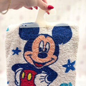May include: White Mickey Mouse towel with a blue border and a smiling Mickey Mouse graphic. The towel features blue stars and a blue and yellow spiral design. The towel is held by a white plastic hanger.