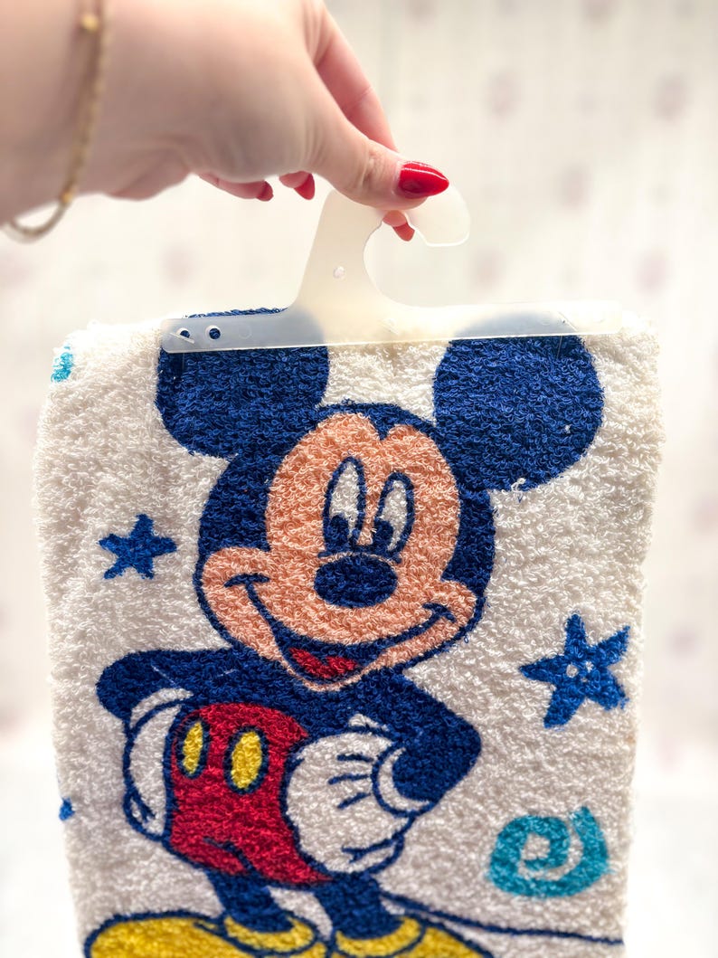May include: White Mickey Mouse towel with a blue border and a smiling Mickey Mouse graphic. The towel features blue stars and a blue and yellow spiral design. The towel is held by a white plastic hanger.