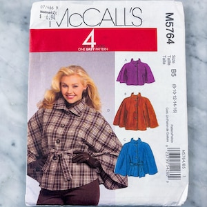 May include: A McCall's M5764 sewing pattern envelope for a cape with four variations. The front shows a person wearing a brown plaid cape with a tie waist, alongside illustrations of purple, orange, and blue capes in sizes 8-10-12-14-16.