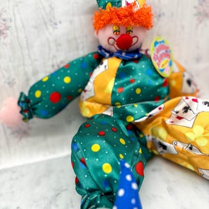 May include: A clown doll with a red nose, orange hair, and a yellow and green hat. The clown's outfit is green with red and yellow polka dots, and yellow with playing card designs. The doll has blue and white polka dot trousers.